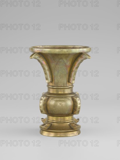 Gu-type vase, Chinese, Qing dynasty 1644–1911, 19th century. Ritual/ceremonial vessel inspired by the cult gu shape with four convex, interrupted strips. Decoration: on the belly, oval reserves with stylized peonies on a grained background. Porcelain/ceramic vessel, height 16 cm, diameter 12.2 cm., A single metallic vessel stands centered against a neutral gray background, displaying a warm golden-bronze color with subtle iridescent patches of green and pink on its surface. The vessel has a flared, trumpet-like upper section with a rounded rim and two curved, stylized protrusions on opposite sides, each showing engraved linear details. The midsection tapers into a rounded, bulbous body decorated with two symmetrical framed panels and faint engraved motifs. The lower portion rests on a stepped, circular base accented by outward-curving, petal-like projections and engraved decorative lines, all sharing the same golden-bronze tone with spots of darker patina.