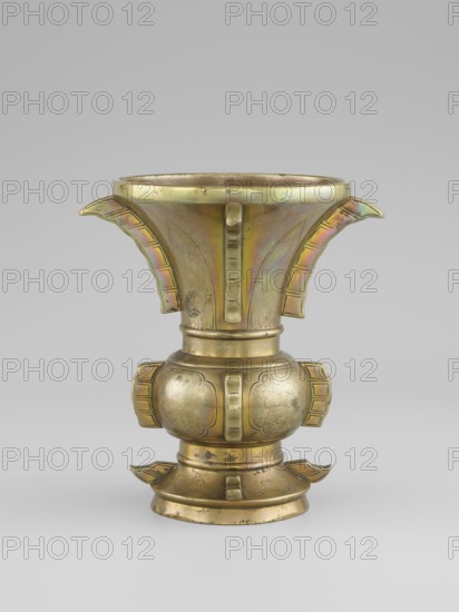 Gu-type vase, noted. Chinese, Qing 1644–1911, 19th century. Ceremonial/ritual vessel inspired by the cult vessel gu with four convex, interrupted strips. Decoration: on the belly, oval reserves with stylized peonies on a grained background. Vessel, height 16 cm, diameter 12.2 cm., A single metallic vessel with a flared top and a rounded midsection sits against a plain light gray background; the vessel shows a few protruding wing-like handles at the top and sides and small decorative tabs around its body, and its surface displays a mix of gold, brass, and subtle iridescent tones with areas of darker patina and slight surface wear.