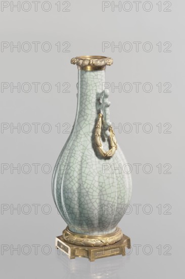 Vase, Chinese, Qing 1644–1911, 19th century. Celadon ceramic slender vase with a long neck and bottle-shaped belly; set on a metal base with the spout framed by a metal ring decorated with acanthus leaves. Height 23.3 cm; diameter 11.5 cm; weight 1.5 kg., A pale celadon vase with a crackled glaze stands on a gold-colored ornate base; the vase has a bulbous body tapering to a narrow neck, a decorative gold-colored rim at the top, and a gold-colored ring attached to a raised sculpted element on one side; visible colors include pale green, gold, and shades of gray in the background and glaze.