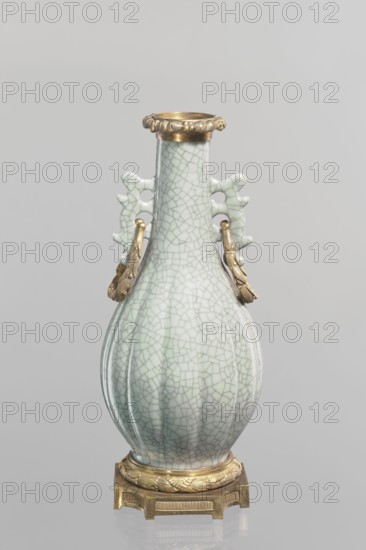 Vase; Chinese; Qing 1644–1911; 19th century. Celadon ceramic decorative vessel — slender vase with long neck and bottle-shaped belly. Set on a metal base; spout framed with a metal ring decorated with acanthus leaves. Height 23.3 cm; diameter 11.5 cm; weight 1.5 kg., A celadon-glazed porcelain vase with a crackle pattern in pale green and off-white sits on a gilt metal base; the vase has a bulbous, vertically fluted body tapering to a narrow neck topped with a decorative gilt rim, and a pair of ornamental pierced handles on either side with attached gilt metal ring fittings that hang down, all set against a neutral gray background.