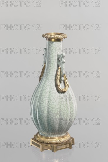 Vase, Chinese, Qing dynasty 1644–1911, 19th century. Celadon ceramic slender vase with long neck and bottle-shaped belly; set on a metal base and the spout framed with a metal ring decorated with acanthus leaves. Height 23.3 cm; diameter 11.5 cm; weight 1.5 kg., A single ceramic vase with a bulbous body and a long neck sits on a metal base; the vase has a pale celadon green glaze with a crackled network pattern and faint darker green lines, and it features decorative applied elements in a metallic gold color including a flared rim, ornamental looped handles on the neck, and a circular foot mount, while the metal base is a darker gold with geometric cutouts; the background is a smooth, light gray.