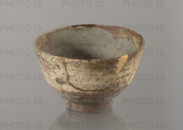 Wan-nari type tea bowl wan-nari chawan, maker, gift; Japanese; Edo period 18th–19th century; stoneware bowl on a low, round foot with rough surface; partly glazed with white milky and grey-bluish glaze with numerous spots and drips; gold lacquer veins; fillings and gluing made with gold lacquer; chawan tea vessel; height 8.4 cm, diameter 13.5 cm., A ceramic bowl with a rounded form and a short foot, showing a weathered surface of off-white, beige, brown, and grey tones with speckled and mottled textures, and visible golden lines filling repaired cracks; the interior is a muted grey-green with spots of brown, the rim shows darker brown wear, and the base has reddish-brown and greenish patina, all set against a neutral grey background with a faint reflection beneath the bowl.