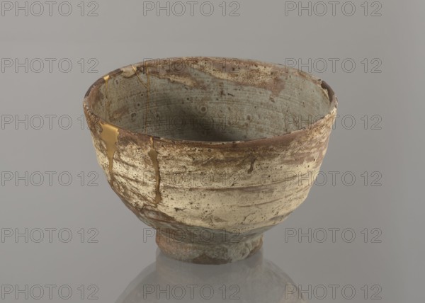 Wan-nari type tea bowl wan-nari chawan, Japanese, Edo 1603–1868, 18th–19th century. Stoneware vessel on a low, round foot; rough surface; partly glazed with white milky and grey-bluish glaze with numerous spots and drips, gold lacquer veins. Fillings and gluing made with gold lacquer. Height 8.4 cm; diameter 13.5 cm., A ceramic bowl sits on a smooth surface against a neutral backdrop, showing an irregular, rounded shape with a slightly flared rim and a narrow foot; the bowl’s exterior and interior surfaces display a weathered texture with mottled patches and speckled markings. The visible colors include muted beige and cream on the exterior, pale blue-gray inside, brown and rusty tones around the rim and base, and a metallic gold filling along repaired cracks. The bowl’s surface shows horizontal striations and areas where glaze appears thin or worn, and the gold repairs create shiny, smooth lines contrasting with the rougher, matte clay. The neutral background is a soft gray, and a fai...
