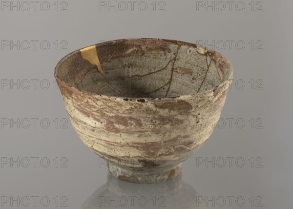 Wan-nari type tea bowl wan-nari chawan, Stoneware bowl on a low, round foot; rough surface; partly glazed with white milky and grey-bluish glaze with numerous spots and drips, with gold lacquer veins. Fillings and gluing made with gold lacquer. Edo period, 18th–19th century, Japan. Function: tea vessel chawan. Height 8.4 cm; diameter 13.5 cm., A weathered ceramic bowl sits against a plain grey background, showing a rough, uneven surface with visible cracks and repairs filled with a metallic gold material. The exterior displays a mottled palette of beige, brown, cream, and muted green with streaks and speckles, while the interior reveals similar tones of pale green, beige, and brown with darker brown lines following the repaired fractures. The rim is irregular and worn, and the base has a slightly darker, aged appearance with the same earthy color variations reflecting faintly on the surface beneath.