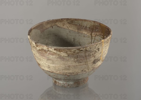 Wan-nari type tea bowl wan-nari chawan, ; Japanese, Edo period 18th–19th century. Stoneware bowl on a low, round foot; rough surface; partly glazed with white milky and grey-bluish glazes with numerous spots and drips, with gold lacquer veins. Fillings and gluing made with gold lacquer. Function: tea vessel chawan. Height 8.4 cm; diameter 13.5 cm; Japan., A ceramic bowl sits on a reflective surface, showing a weathered, uneven texture with visible cracks and repaired seams. The bowl's exterior and interior display muted tones of beige, cream, light brown, and gray with areas of darker brown speckling and mottling. A glossy golden-colored material fills and runs along some of the cracks. The rim is irregular and slightly chipped in places. The background is a soft, uniform gray.