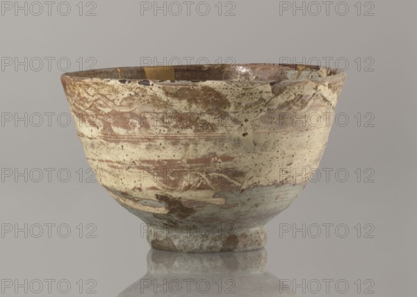 Wan-nari type tea bowl wan-nari chawan, Stoneware bowl on a low, round foot; rough surface; partly glazed with white milky and grey-bluish glaze with numerous spots and drips, with gold lacquer veins. Fillings and gluing made with gold lacquer. Edo period, 18th–19th century, Japan. Function: tea vessel chawan. Height 8.4 cm; diameter 13.5 cm., A shallow, rounded ceramic bowl sits on a reflective surface, showing a textured, weathered exterior with uneven glazing. The bowl displays a mix of beige, cream, brown, and tan tones with areas of muted greenish-blue near the base and subtle darker brown spots and streaks. The rim appears slightly irregular and chipped in places, and there is a visible repaired area with a metallic gold patch along the inner edge. The surface shows mottled speckling and faint horizontal decorative bands, and the interior has a glossy finish in shades of brown and cream.
