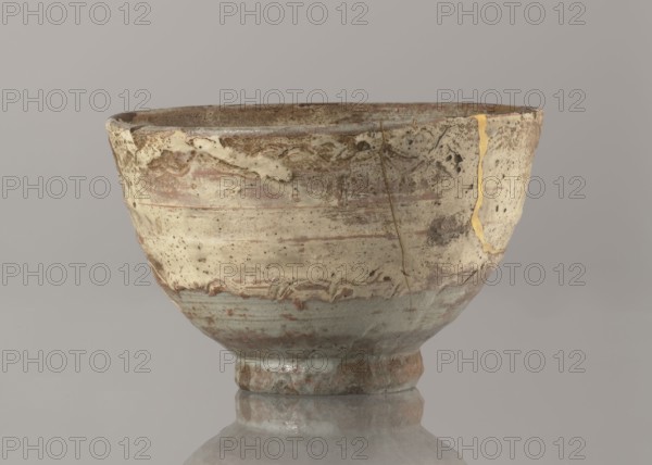 Wan-nari type tea bowl wan-nari chawan, Bowl on a low, round foot; rough surface; stoneware partly glazed with white milky and grey-bluish glaze with numerous spots and drips, gold lacquer veins. Fillings and gluing made with gold lacquer. Edo period, 18th–19th century Japan. Function: tea vessel chawan. Height 8.4 cm; diameter 13.5 cm., A single ceramic bowl sits centered on a reflective surface, showing a worn and irregular rim and an overall rounded shape that narrows to a small foot. The surface of the bowl displays layered patches of muted cream, beige, light brown, and gray with areas of darker brown spotting and smudging; there are visible thin cracks repaired with a metallic gold-colored material and faint streaks and bands of reddish-brown and pale blue-green glaze near the lower portion. The interior rim appears darker and more textured while the exterior shows a mix of smooth glazed areas and rough, pitted sections, and the reflection beneath the bowl echoes the same muted c...