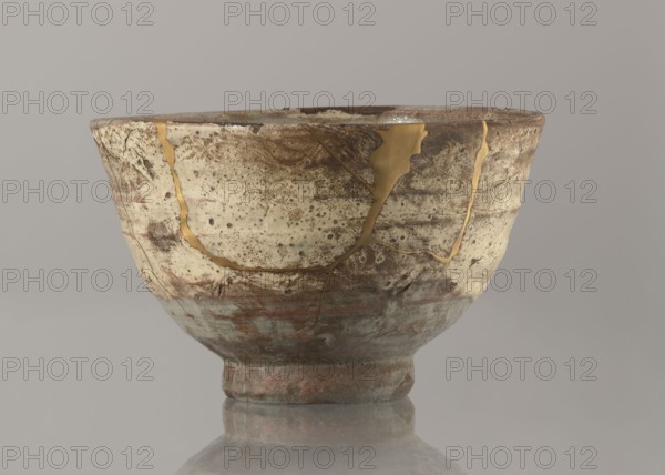 Wan-nari type tea bowl wan-nari chawan, bowl on a low, round foot; rough surface; stoneware partly glazed with white milky and grey-bluish glaze with numerous spots and drips, gold lacquer veins. Fillings and gluing made with gold lacquer. 18th–19th century Edo period, Japan. Height 8.4 cm; diameter 13.5 cm. Japanese art; ceramics; chawan tea vessel., A ceramic bowl with a rounded body and a narrow foot sits on a reflective surface; its surface shows a mottled, weathered texture with areas of beige, off-white, brown, rust, and muted greenish-blue, and visible repaired cracks filled with a shiny golden material that form irregular lines and patches across the exterior.