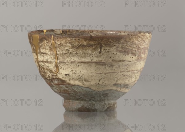 Wan-nari type tea bowl wan-nari chawan, noted. Japanese, Edo period 18th–19th century. Stoneware bowl on a low, round foot; rough surface; partly glazed with white milky and grey-bluish glaze with numerous spots and drips and gold lacquer veins. Fillings and gluing made with gold lacquer. Function: tea vessel chawan. Height 8.4 cm; diameter 13.5 cm., A ceramic bowl with a worn, textured surface sits against a plain background, showing a palette of beige, brown, tan, gray, blue-gray, and gold. The bowl has an uneven rim and mottled areas where darker brown and rust tones blend into lighter beige and tan, with speckled dark spots and subtle horizontal striations. A few gold-colored repairs trace irregular lines along the side and rim. The lower portion shifts to cooler gray and blue-gray hues with rougher texture, and the bowl rests on a short, slightly irregular foot that mirrors the same earthy tones. The background is a smooth neutral gray.