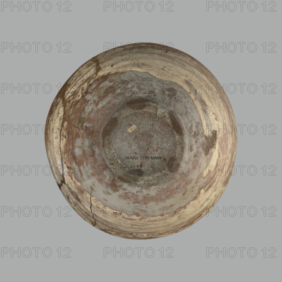 Wan-nari type tea bowl wan-nari chawan, gift; Japanese art, Japanese, Edo 1603–1868; stoneware chawan tea vessel on a low, round foot; rough surface; partly glazed with white milky and grey-bluish glaze with numerous spots and drips, with gold lacquer veins. Fillings and gluing made with gold lacquer. 18th–19th century Edo period, Japan. Height 8.4 cm; diam. 13.5 cm., A top-down view of a circular, shallow bowl with a weathered, uneven surface showing a mix of gray, beige, brown, and rust-red tones, with areas of mottled discoloration and patches of lighter cream, a few hairline cracks and darker smudges around the rim, and a small rectangular sticker near the center, all set against a plain gray background.