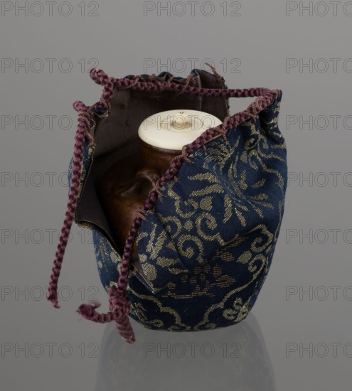 Tea canister chaire of the mimitsuki type. Brown container with two handles and a lid in a navy blue brocade bag silk. Dark brown, porous clay, wheel-thrown; light brown glaze with rough air bubble cracks; applied plastic handles. 19th century late Edo–Meiji period, Owari, Japan. Height 6.3 cm; diameter 5 cm. Function: tea container chaire., A fabric pouch with a drawstring closure sits open to reveal a rounded container inside; the pouch fabric is predominantly deep blue with a metallic gold floral and scroll pattern and has a dark interior lining, while the drawstring is a braided purple cord with frayed ends; the container inside appears brown with a glossy surface and a creamy off-white lid with a small raised knob; the background is a smooth neutral gray and the objects cast a subtle reflection on the surface below.