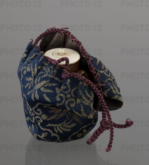 Tea canister chaire of the mimitsuki type, Seto; Japanese, Edo 1603–1868 / Meiji 1868–1912, 19th century; brown container with two handles and a lid in a navy blue silk brocade bag; dark brown porous wheel-thrown clay with light brown glaze showing rough air-bubble cracks; plastic handles; height 6.3 cm, diameter 5 cm; Owari, Japan., A fabric pouch with a blue background patterned in gold sits open, revealing a light-colored circular lid and a glimpse of a brown rounded object inside; the pouch has braided drawstring cords in a purple tone that form a knot and hang down, and the pouch interior appears darker, while the overall scene rests against a neutral gray surface and background.