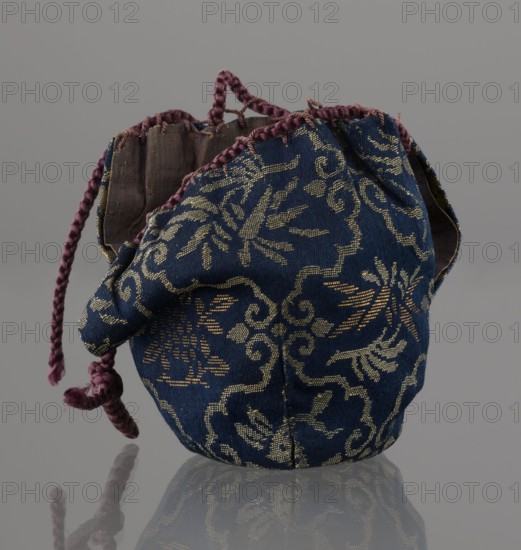 Tea canister chaire of the mimitsuki type, Seto. Brown container with two handles and a lid in a navy blue brocade bag silk. Dark brown, porous clay, wheel-thrown; light brown glaze with rough air bubble cracks; plastic handles. 19th century Edo/Meiji period, Japan, Owari. Vessel chaire, height 6.3 cm, diameter 5 cm., A small fabric drawstring pouch with a blue exterior patterned in metallic gold designs and a brownish interior, edged and gathered with braided purple cords and a knotted purple tie, shown against a neutral gray background with a soft reflection on the surface.