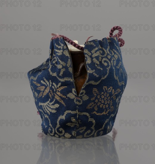 Tea canister chaire of the mimitsuki type. Brown container with two handles and a lid in a navy blue brocade bag silk. Dark brown, porous clay, wheel-thrown; light brown glaze with rough air bubble cracks; plastic handles. 19th century late Edo–Meiji period, Owari, Japan. Vessel; height 6.3 cm, diameter 5 cm., A small fabric pouch with a patterned woven surface in deep blue and metallic gold tones, showing a floral and scrolling motif, is pictured against a neutral gray background; the pouch is slightly open at the top revealing a glimpse of a brown ceramic object inside and a small portion of an off-white rim, and a braided cord in muted purple emerges from the opening and loops at the top, with the pouch resting on a reflective surface that mirrors its colors and shapes.