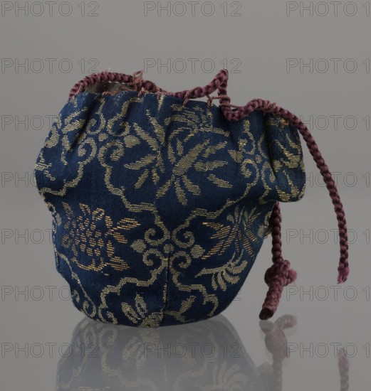Tea canister chaire of the mimitsuki type. Brown container with two handles and a lid in a navy blue brocade bag silk. Dark brown, porous clay, wheel-thrown; light brown glaze, rough air bubble cracks, plastic handles. 19th century Edo period / Meiji period, Japan, Owari. Vessel. Height 6.3 cm; diameter 5 cm. Ceramics; containers; chaire vessel; tea., A small drawstring pouch made of deep blue fabric with an ornate woven pattern in a metallic gold color, showing floral and scrolling motifs; the pouch has a gathered top finished with a braided cord in a muted purple color that extends out and loops with a small knot at the end; the fabric shows slight creasing and varied texture, and the pouch rests on a reflective gray surface with a neutral gray background, the reflection of the pouch and purple cord visible on the surface.