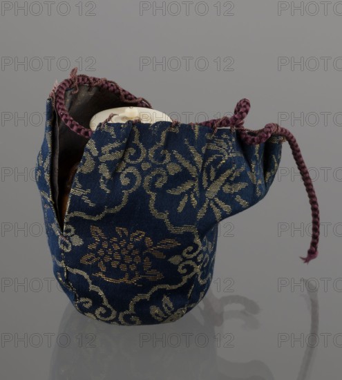 Tea canister chaire of the mimitsuki type, Seto; Japanese, Edo 1603–1868–Meiji 1868–1912; 19th century; brown container with two handles and a lid in a navy blue brocade silk bag; dark brown porous wheel-thrown clay with light brown glaze showing rough air-bubble cracks; plastic handles; Owari, Japan; vessel; height 6.3 cm, diameter 5 cm., A small fabric pouch with a drawstring sits on a smooth reflective surface; the pouch is primarily deep blue with an ornate woven pattern in a gold tone, and it has a braided drawstring that is a muted purple color; the pouch opening reveals a glimpse of a round, off-white object inside and the interior lining appears dark brown; the reflection of the pouch and drawstring is visible on the grayish surface beneath it.