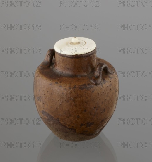 Tea canister chaire of the mimitsuki type. Brown container with two handles and a lid in a navy blue brocade bag silk. Dark brown, porous clay, wheel-thrown; light brown glaze with rough air bubble cracks; applied handles. 19th century late Edo / Meiji period, Owari, Japan. Height 6.3 cm; diameter 5 cm., A small rounded ceramic jar with a short neck and a pair of looped handles, displaying a mottled brown surface with darker brown spots and areas of rougher texture, topped by a smooth off-white circular lid with a small raised knob, all set against a plain gray background with a faint reflective gray surface beneath.
