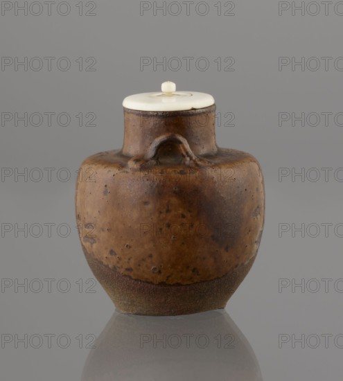 Tea canister chaire of the mimitsuki type, Seto; . Japanese art; Edo 1603–1868 / Meiji 1868–1912. Ceramics container chaire for tea. Brown container with two handles and a lid in a navy blue brocade bag silk. Dark brown porous wheel-thrown clay; light brown glaze with rough air bubble cracks; applied handles. 19th century. Japan, Owari. Vessel; height 6.3 cm, diameter 5 cm., A rounded ceramic jar with a short neck and a small handle-like protrusion near the top sits centered against a plain background. The jar shows a mottled surface in shades of brown and darker brown with some speckled spots and a rougher textured lower band in darker earthy brown. A pale, cream-colored circular lid with a small knob rests on the neck of the jar. The background and surface beneath the jar are gray, and a subtle reflection of the jar appears below it.
