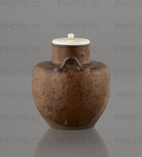 Tea canister chaire of the mimitsuki type, Seto; gift; Japanese art; Japanese culture; Edo 1603–1868 — Meiji 1868–1912; ceramics; container chaire, tea. Brown container with two handles and a lid in a navy blue brocade bag silk. Dark brown, porous wheel-thrown clay; light brown glaze with rough air bubble cracks; plastic handles. 19th century; Japan, Owari. Vessel; height 6.3 cm; diameter 5 cm., A rounded ceramic jar with a short neck and a small arched handle near the top sits centered against a plain background; the jar's surface is mottled brown with darker speckles and patches and a slightly rough texture, and it has a lighter tan area near the base; a smooth, pale lid rests on the neck with a small pale knob on top, and the jar casts a faint reflection on the glossy surface beneath; visible colors include brown, tan, pale off-white, gray, and hints of darker brown.