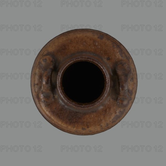 Tea canister chaire of the mimitsuki type. Brown container with two handles and a lid in a navy blue brocade bag silk. Dark brown, porous wheel-thrown clay; light brown glaze with rough air bubble cracks; applied plastic handles. 19th century Edo–Meiji period, Owari, Japan. Vessel chaire for tea, height 6.3 cm, diameter 5 cm., A round, top-down view of a brown, weathered ceramic vessel with a central dark opening and a textured rim, showing a few raised, handle-like protrusions on opposite sides; the vessel surface has mottled shades of brown with darker spots and slight variations, and it sits against a flat gray background.