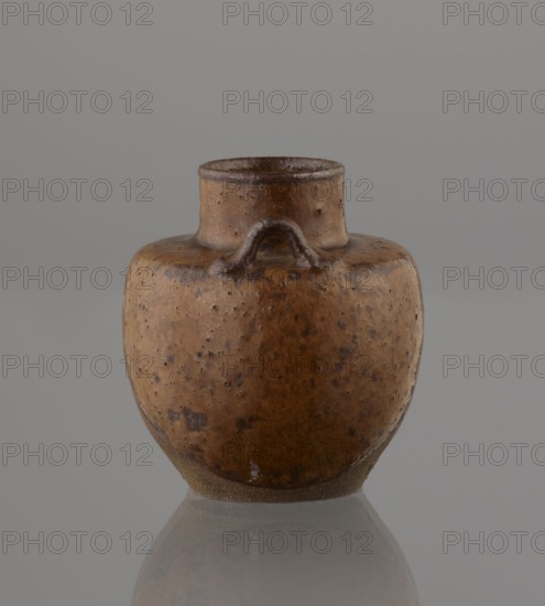 Tea canister chaire of the mimitsuki type, Japanese, Edo 1603–1868 / Meiji 1868–1912, 19th century. Brown container with two handles and a lid, in a navy blue brocade silk bag. Dark brown, porous wheel-thrown clay with light brown glaze showing rough air-bubble cracks; plastic handles. Owari, Japan. Height 6.3 cm; diameter 5 cm., A ceramic vessel with a rounded body and a short cylindrical neck sits centered against a plain background, showing a textured surface with small pits and irregular mottling; its colors include brown, tan, and darker brown spots with some subtle darker areas around the neck and handle ridge, and a slightly lighter tan band near the base, while a muted gray background and a faint reflection below are also visible.