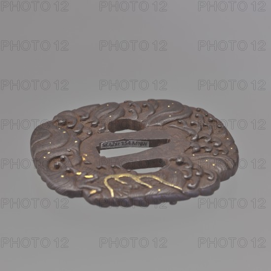 Crossguard tsuba, Kikuchi Tsunekatsu 1704-? maker; Tsuba in the shape of a mokkô quatrefoil with seppa dai oval. Decorated on both sides with a high-relief image of a dragon among rough waves; creature's claws and horns inlaid with gold using the nunome technique; tiny gold dots illuminate the waves. Signed. 1st half of the 18th century Edo period, 1603–1868, Japan. Materials: metal with gold inlay. Dimensions: height 7.9 cm, width 7.1 cm, depth 0.4 cm. Subjects: sword crossguard, katana, dragons, sea waves., A round, ornate metal object with carved, swirling raised patterns and rounded studs, showing a dark brown and rust color overall with small areas of gold-colored accents; it has an oval central opening and two elongated slots, and the background is a smooth light gray.