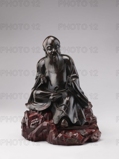 Sage sitting on a rock base;, Chinese art; Chinese culture; Qing 1644–1911; figurine of a man—scholar/old man associated with Chinese Buddhism; 18th–19th century; China; sculpture; height 32.5 cm., A dark brown wooden statue of a seated robed figure with a long beard and a smooth rounded head sits on an irregular carved base that is reddish-brown; the figure’s garments have flowing folds and polished highlights, with one hand resting on a knee and the other holding a small object, and the base is textured with angular, rock-like forms and scattered rounded elements, all set against a pale gray background.