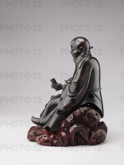 Sage sitting on a rock base, unknown sculptor, Chinese art, Chinese culture, Qing 1644–1911, figurine of a man, Chinese Buddhism, scholar/old man, 18th–19th century, China, sculpture, height 32.5 cm, A carved figurine of a seated elderly man with a long beard, shown in profile, carved in a dark brown to near-black material; he wears flowing robes with smooth curved folds and holds a small rectangular object in his hands; the figure sits on a textured base resembling rock or stylized cloud forms in reddish-brown with darker and lighter patches; the background is a smooth neutral gray fading lighter toward the bottom.