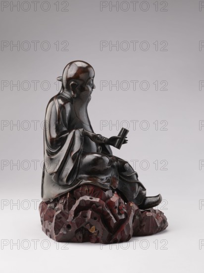 Sage sitting on a rock base; sculptor, Chinese art; Chinese culture; Qing 1644–1911; figurine of a man—scholar/old man associated with Chinese Buddhism; 18th–19th century, China; sculpture; height 32.5 cm., A dark brown wooden sculpture of a seated elderly figure with a long beard, wearing flowing robes and holding a small rectangular object, positioned on a textured reddish-brown rocky base, set against a light gray background; visible colors include dark brown, reddish-brown, black, and light gray.