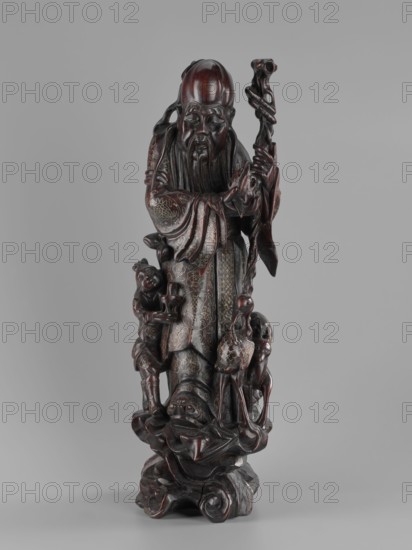 Deity of Longevity Shouxing/Shoulao holds a long traveling staff and a peach of immortality; his robe is decorated with cloud motifs, lingzhi mushrooms, and cranes in medallions. At his feet stand a servant with a vessel, a deer, and a crane. Figurine sculpture in Chinese art with peaches, cranes, and other animal iconography. Qing dynasty 1644–1911, 19th century, height 54.2 cm., A carved dark brown wooden figurine of an elderly man with a long beard and a high forehead stands holding a tall twisted staff; he wears flowing robes with carved patterned details and stands on a sculpted base with organic, wave-like forms, accompanied by a few small figures and animal-like shapes clustered around his legs; the surface shows polished highlights and deeper reddish-brown tones against a neutral gray background, with areas of lighter brown and subtle darker accents across the carving.