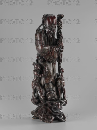 Deity of Longevity Shouxing/Shoulao holds a long traveling staff and a peach of immortality. His robe is decorated with cloud motifs, lingzhi mushrooms, and cranes in medallions. At his feet stand a servant holding a vessel, a deer, and a crane. Peaches, figurines, cranes, and other animals feature in the iconography. Sculptural work in Chinese art style, Qing dynasty 1644–1911, 19th century, height 54.2 cm., A tall carved figure rendered in dark brown and deep reddish-brown tones stands on a sculpted base of swirling forms, with carved drapery and textured patterns on the clothing, a long beard, and a hand holding a tall, twisted staff; a smaller carved figure and an animal-like figure are positioned near the base, and the whole piece shows polished highlights and darker recesses against a plain light gray background.