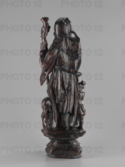 Deity of Longevity Shouxing/Shoulao holds a long traveling staff and a peach of immortality; his robe is decorated with cloud motifs, lingzhi mushrooms, and cranes in medallions. At his feet stand a servant with a vessel, a deer, and a crane. Stone/ceramic sculpture with peaches, figurines, cranes, and animals; Chinese, Qing dynasty 1644–1911, 19th century, height 54.2 cm., A carved wooden figure viewed from the back showing a robed person standing on a faceted base, holding a gnarled staff in one hand; a smaller figure stands beside the larger figure and an animal with a curved neck is positioned near the base; the carving has a deep reddish-brown wood tone with darker brown areas and small lighter-colored decorative inlays or spots, and the background is a uniform pale gray.