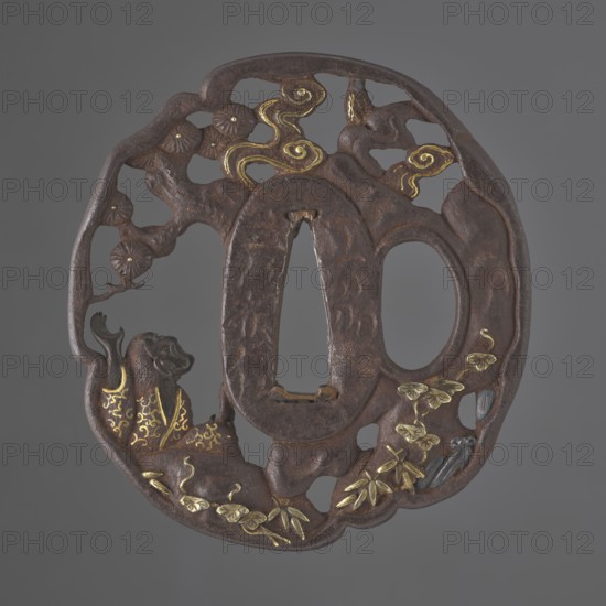 Crossguard tsuba, Sôten; Kitagawa Sôten attributed. Tsuba oval tatemaru gata with an irregular, rounded edge, decorated with positive openwork, inlaid with gold and shakudo, depicting Uba under a pine tree by a stream looking at the sky as the moon emerges from behind clouds. Signed. Materials: gold, shakudo. Subject: pines, bamboos, plants, waterfall. Function: sword crossguard for katana. Date: 1701–1730 Edo period, 1603–1868. Place: Japan; Ômi Eshu province. Dimensions: height 7.1 cm, width 6.7 cm., A round, openwork metal object with a dark brown, weathered surface and patches of golden inlay depicting swirling cloud-like shapes and stylized leaves; the piece has an irregular outer rim and an oval central opening with a second smaller opening beside it. A sculpted figure with a raised arm and a carved head appears on one side, highlighted with gold decorative patterns on its clothing. Branch-like forms with round floral elements extend across the top left, and a cluster of small, g...