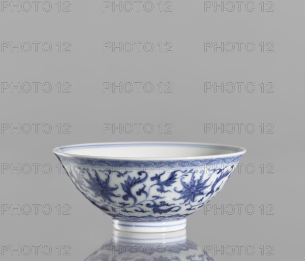 Bowl, blue-and-white porcelain, China, Qianlong style, Qing dynasty 1644–1911, 1736–1795. Small vessel with slightly flared rim decorated with lotuses and lotus tendril ornament. Height 4.3 cm; diameter 9.8 cm., A ceramic bowl with a white base and blue floral and vine patterns along its exterior, sitting on a short foot and reflecting faintly on the surface below; the background is a uniform light gray.