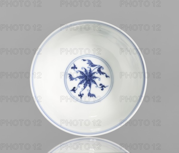 Bowl, blue and white porcelain, Chinese, Qianlong style, Qing dynasty 1644–1911, 1736–1795. Porcelain vessel with lotus and lotus tendril ornament; slightly flared bowl. Height 4.3 cm; diameter 9.8 cm., A white ceramic bowl with a smooth glossy surface sits against a gray background, showing a delicate blue floral-like motif painted at the center inside a thin blue circular border, and thin blue lines rim the inner edge; the decoration includes flowing petal and vine shapes in blue, and the bowl's interior and exterior appear predominantly white with highlights and reflections of light, while the background is gray.