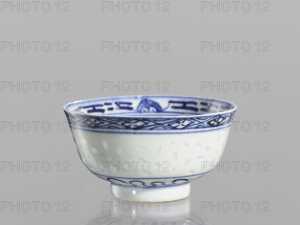 Bowl, unknown provenance; Chinese; Qing dynasty 1644–1911; ceramics, blue-and-white porcelain. Bowl on a low ring foot, decorated on the body with a rice grain rice pattern ornament and geometric-plant borders; in the mirror a lotus flower in a double circle. Floral and plant ornaments, lotuses. 18th–19th century, China; vessel; height 5.5 cm, diameter 10.3 cm., A small porcelain bowl sits against a plain light gray background, showing a rounded body with a short foot. The bowl is primarily white with blue decorative bands and motifs: a dark blue patterned band near the rim featuring interlaced geometric shapes, a thinner blue line beneath it, and a blue wave-like band near the foot. The interior rim also displays blue decorative elements. The white body has a subtle raised leaf-like pattern visible as pale bluish-green shapes against the white surface. The bowl casts a soft reflection on the glossy surface below, and the visible colors are white, several shades of blue, pale bluish-gr...