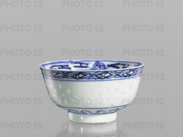 Bowl, unknown provenance; Chinese; Qing dynasty 1644–1911; blue-and-white porcelain; rice pattern; geometric and plant ornaments; floral decorations with lotuses; bowl on a low ring foot; body decorated with a rice grain ornament and geometric-plant borders; in the mirror a lotus flower in a double circle; 18th–19th century; China; vessel; height 5.5 cm, diameter 10.3 cm., A small ceramic bowl sits against a plain gray background, showing a white base color with blue decorative patterns. The rim area features a dark blue geometric border and additional blue motifs visible inside the bowl, while the body displays a subtle pattern of pale blue leaf-like shapes. The foot of the bowl has a thin blue line and a wavy blue band above it, and the bowl’s glossy surface produces soft reflections on the surface below.