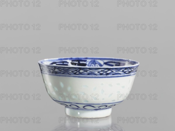 Bowl, Chinese, Qing 1644–1911, ceramics, blue-and-white porcelain. Rice-pattern and geometric-plant ornaments with floral decorations and lotuses. Bowl on a low ring foot, decorated on the body with a rice grain ornament and geometric-plant borders; in the mirror a lotus flower in a double circle. 18th–19th century, China. Vessel; height 5.5 cm, diameter 10.3 cm., A small ceramic bowl with a white base and blue decoration, showing a band of intricate blue geometric and floral patterns around the rim and a simpler blue band near the foot; the bowl has a glossy finish with subtle pale blue leaf-like motifs on the white surface and sits on a short circular foot against a neutral gray background.