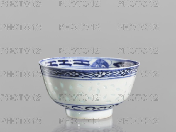 Bowl, Chinese, Qing 1644–1911, ceramics; blue-and-white porcelain with rice-pattern and geometric and plant/ floral lotus decorations. Bowl on a low ring foot, decorated on the body with a rice grain ornament and geometric-plant borders; in the mirror a lotus flower in a double circle. 18th–19th century, China; vessel; height 5.5 cm, diameter 10.3 cm., A ceramic bowl with a glossy white surface and blue decorative patterns around the rim and base sits on a reflective surface; the rim features a darker blue band with geometric motifs and a thinner blue line below it, the interior rim also shows blue designs, and the bowl’s body displays a subtle pattern of small, pale leaf-like shapes against the white glaze, with the visible colors being white, several shades of blue, and a hint of pale green from reflections, all set against a plain light gray background.