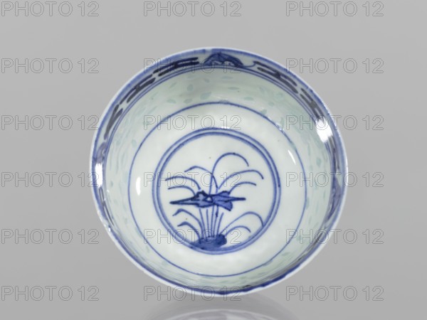 Bowl, Chinese, Qing dynasty 1644–1911, 18th–19th century. Blue-and-white porcelain, rice pattern with geometric and plant ornaments and floral decorations including lotuses. Bowl on a low ring foot, decorated on the body with a rice grain ornament and geometric-plant borders; in the mirror a lotus flower in a double circle. Vessel; height 5.5 cm, diameter 10.3 cm., A round ceramic bowl viewed from above with a pale white interior and blue painted decoration, showing a central circular motif with stylized plant-like lines and curved shapes in blue, concentric blue rings around the center, a patterned blue band near the rim, and a few light green brushstroke spots on the inner walls; the rim and painted details appear in shades of blue against the white background, and the overall scene is set on a neutral gray surface.