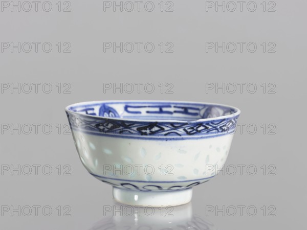 Bowl, Chinese, Qing dynasty 1644–1911, blue-and-white porcelain. Bowl on a low ring foot, decorated on the body with a rice grain rice pattern ornament and geometric and plant borders; in the central mirror a lotus flower within a double circle. 18th–19th century, China. Vessel; height 5.3 cm, diameter 10.3 cm., A small ceramic bowl with a rounded shape and a short foot, primarily white with blue decorative patterns; the rim features a band of dark blue geometric and scroll-like designs, the interior rim also showing blue motifs, and the lower exterior has a lighter blue band with wave-like patterns and scattered pale blue teardrop shapes on the white surface; the background is a smooth light gray and the bowl casts a faint reflection on the surface beneath it.