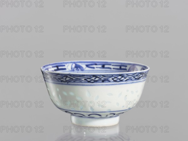 Bowl, Chinese, Qing dynasty 1644–1911, 18th–19th century. Blue-and-white porcelain bowl on a low ring foot, height 5.3 cm, diameter 10.3 cm. Decorated on the body with a rice grain pattern, geometric and plant borders, floral decorations including lotuses; in the mirror a lotus flower within a double circle., A small ceramic bowl sits against a neutral gray background, showing a glossy white surface with blue decorative bands and patterns around the rim and base; the rim features a darker blue geometric border and interior motifs in blue, while the body displays pale blue leaf-like spots and a thin blue line near the foot.