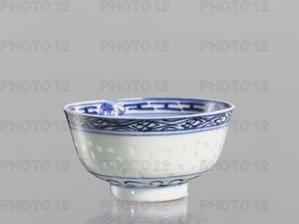Bowl, Chinese, Qing dynasty 1644–1911, 18th–19th century. Blue-and-white porcelain bowl on a low ring foot, height 5.3 cm, diameter 10.3 cm. Decorated on the body with a rice grain rice pattern ornament, geometric and plant borders, floral decorations including lotuses; in the mirror a lotus flower in a double circle., A small porcelain bowl with a rounded shape and a short foot, predominantly white with blue decorative patterns; the rim and inner band feature darker blue geometric and interlaced designs, while the exterior body shows a softer blue dotted or leaf-like motif and a thin blue line near the base; the bowl sits on a reflective surface and the background is a smooth light gray.
