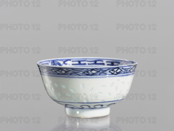 Bowl, unknown provenance; Chinese; Qing dynasty 1644–1911; blue-and-white porcelain; 18th–19th century; vessel on a low ring foot, height 5.3 cm, diameter 10.3 cm; decorated on the body with a rice grain pattern, geometric and plant borders; mirror with a lotus flower in a double circle; motifs include rice pattern, geometric ornaments, plant ornaments, floral decorations, lotuses., A small ceramic bowl sits on a reflective surface against a plain gray background, showing white and blue as the dominant colors; the bowl has a white body with blue decorative bands near the rim and base, a patterned blue border around the upper interior and exterior, and a subtle light blue leaf-like motif scattered on the midsection; the rim is slightly undulating and the bowl rests on a short circular foot with a blue line near its top, and soft reflections and highlights are visible on the glazed surface.