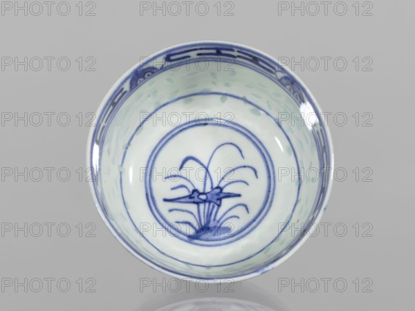 Bowl, Chinese, Qing dynasty 1644–1911, ceramics; blue-and-white porcelain with rice-pattern ornament, geometric and plant borders, floral decorations and lotus motif in the central mirror within a double circle. Bowl on a low ring foot. 18th–19th century, China. Height 5.3 cm; diameter 10.3 cm., A round ceramic bowl viewed from above with a glossy white interior and blue painted decoration; the rim has a band of darker blue patterning, the interior sides show a few pale green brush marks, and the center contains a blue circular border enclosing a stylized blue plant-like motif.