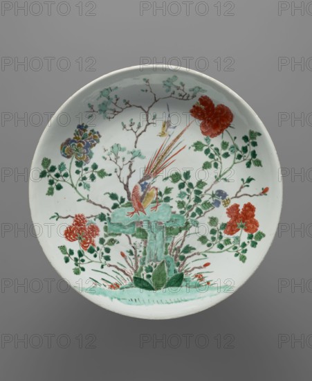 Platter, pheasants, flowers, colored porcelain, Qing style, Chinese art, Chinese culture, Qing 1644–1911, 18th–19th century Qing dynasty, China, vessel; height 5.9 cm, diameter 35.3 cm, A round ceramic dish with a pale white background decorated in green, red, blue, yellow, and brown pigments shows a central scene of a stylized stone pedestal rendered in green with leafy plants at its base, surrounded by flowering branches and foliage. Perched on the pedestal is a colorful bird with a long tail featuring red and yellow accents and blue markings on its body, while a smaller bird in flight appears nearby. Around the main motif are a few clusters of large red flowers, several sprays of green leaves, and a multicolored bloom with blue and yellow tones, all connected by slender brown branches and twigs against the pale surface.