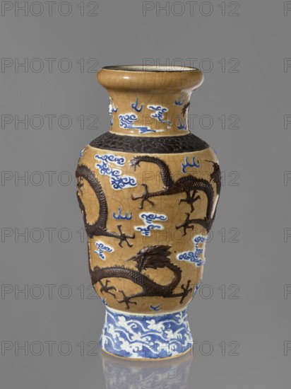 Vase, Chinese, 19th century. Steam vase; ovoid; short, slightly flared foot; pear-shaped belly; neck bulging at the rim. Ceramic vessel decorated with dragons, clouds, flames, cracklework, and geometric ornaments. Height 43.6 cm; diameter 15 cm., A ceramic vase with a glossy finish stands against a plain gray background, showing a warm tan body with a crackled surface, dark brown raised dragon motifs winding around the midsection, and several decorative cloud-like shapes in blue and white scattered across the tan areas; a band near the shoulder features a textured dark brown pattern, and the flared neck shares the tan color with blue and white cloud shapes, while the foot of the vase contrasts in blue and white with a swirling wave pattern.