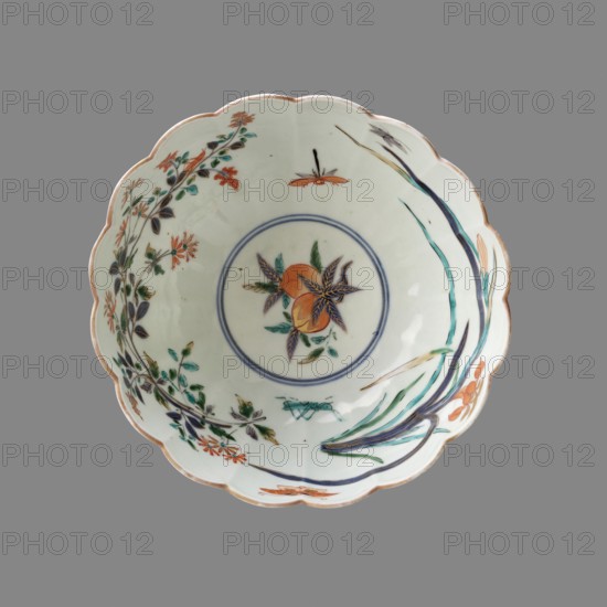 Bowl, Japanese, Edo 1603–1868, late 17th century Edo period, Arita, porcelain Imari, vessel, height 12.0 cm, diameter 20.5 cm; decoration: floral pattern, figural scenes, peaches, dragonflies, crickets., A scalloped ceramic bowl with a pale off-white interior decorated with painted botanical motifs, featuring a central circular medallion containing a stylized fruit and leaves in orange, green, and dark blue, surrounded by a thin double ring in muted blue; the inner surface shows a spray of slender green and teal grasslike leaves and stems with orange flowers, and a curved arrangement of leafy branches in shades of green, teal, dark blue, and orange with small orange blossoms along the rim; the rim has a thin brownish edge and the background behind the bowl is a flat medium gray.
