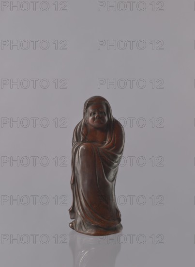Daruma, unknown sculptor, Japanese, Meiji period 1868–1912, ceramics figurine of a male Buddhist monk Daruma wearing headgear and a long, flowing robe covering the head, late 19th century, Seto, Japan; height 21.2 cm, A small carved figure made of dark brown material stands centered against a smooth light gray background, showing a robed person with a rounded head and a solemn facial expression, draped garments with flowing folds that wrap around the body, subtle highlights and deeper brown shadows on the surface, and a glossy finish that creates a faint reflection beneath the base.