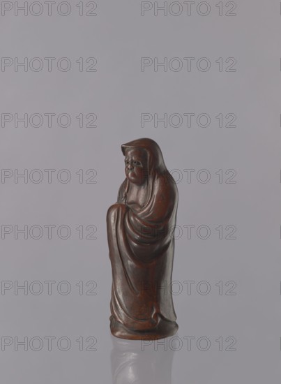 Daruma, Japanese, Meiji period 1868–1912. Ceramic figurine of Daruma Buddhist monk, male figure in a long flowing robe covering the head and headgear. Late 19th century, Seto, Japan. Height: 21.2 cm., A small carved figurine with a rounded head and draped cloak, standing upright on a smooth base, showing curved folds and rounded contours; the surface is polished and appears dark brown with lighter brown highlights, set against a pale gray background with a subtle reflection of the base on a glossy surface.